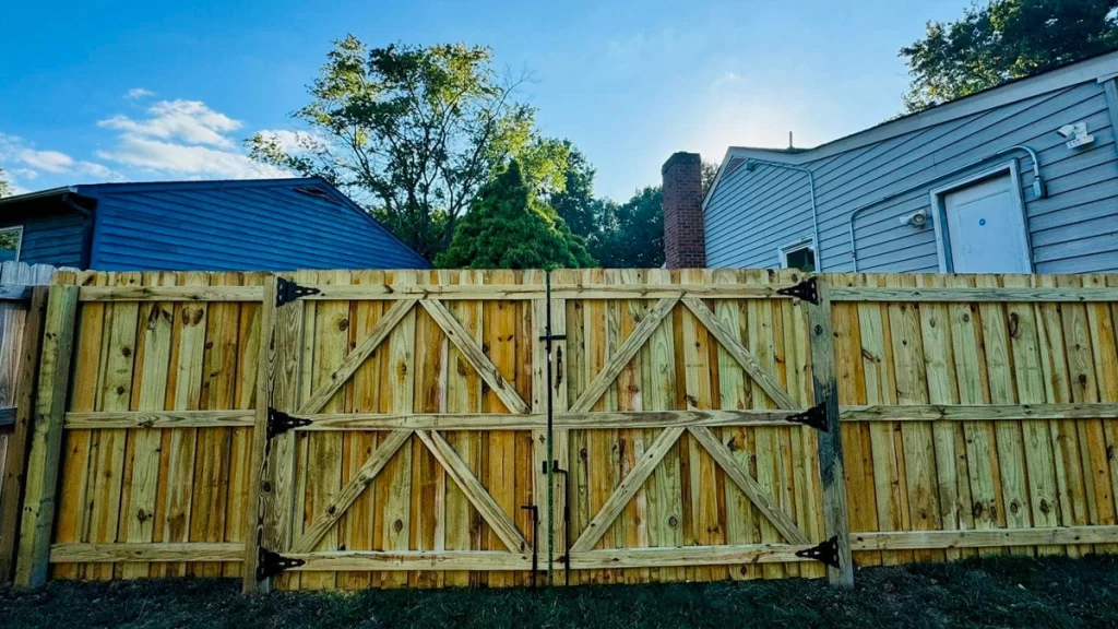 A wooden privacy fence with double gates featuring decorative black hardware and a cross-brace design, enclosing a residential backyard. Choosing the perfect fence begins with deciding what the fence should do. 