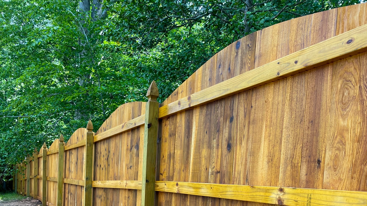 A beautifully crafted wood privacy fence with a scalloped top design and finials on the posts, set against a lush green background. First Step Fence offers custom residential wood fence design and installation services to enhance privacy, security, and curb appeal in Richmond, Virginia, and the surrounding areas.