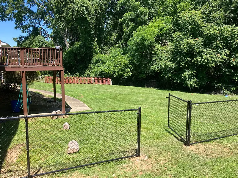 A black, vinyl-coated chain link fence with an open single gate on a residential property. First Step Fence install chain link fences and gates for homeowners in Richmond, VA and the surrounding areas.